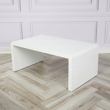 An angled shot of a white coffee table on a background of white brick, and laminate flooring, to demonstrate how the product may look in a home
